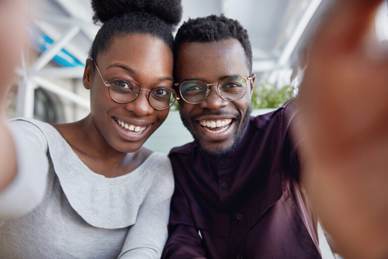 Glad African friends enjoy resting together, have positive expressions, make photo, stretch hand as take selfie, share pictures in social networks. African American woman and man in spectacles